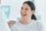 A woman smiles at a person in a white coat touching her shoulder. Bright, airy setting with blurred background and soft colors, conveying joy.