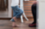Child in blue jeans stands on tiptoes facing seated adult in red shoes. Indoor wood floor, relaxed setting.