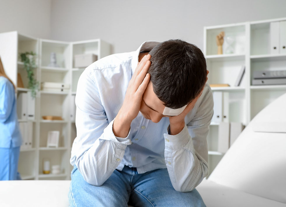 Man with bandaged forehead sitting and holding his head in pain, a nurse in blue uniform in the background, white shelves behind.