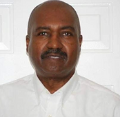 A man is standing in front of a white door smiling. He has a white shirt and a black mustache.