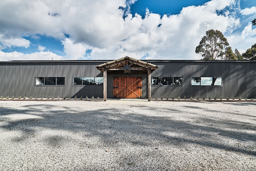 A large grey modern shed with wooden, barn style doors and a portico made with recycled timber. Placed in front of a bed of white rock, with nature in the background.