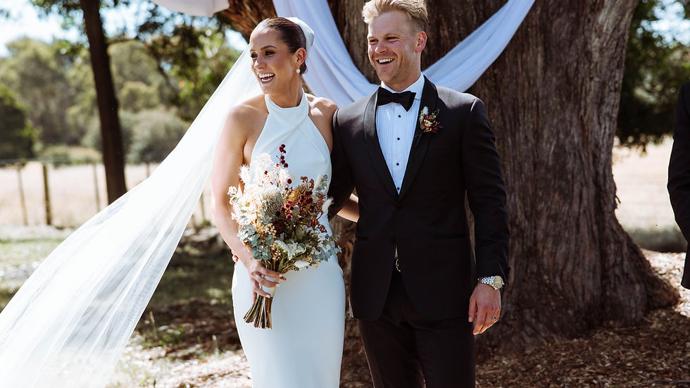 Bride in white gown holds bouquet, groom in black suit smiles. They're standing by a tree draped in white fabric, outdoors in sunlight.
