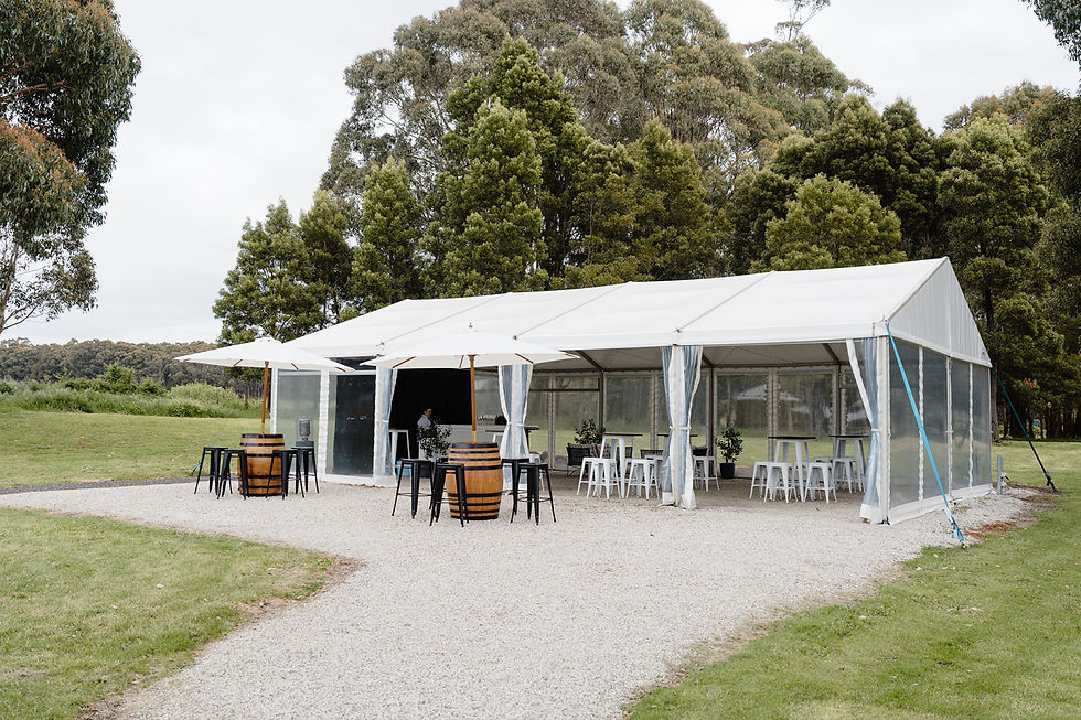 White event tent with tables and stools, set in a lush green park. Two umbrellas and barrel tables add charm. Overcast sky above.