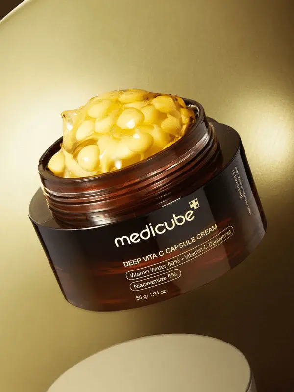 Brown jar of Medicube Deep Vita C Capsule Cream with yellow capsules inside, against a gold background. Text on jar highlights ingredients.