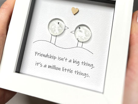 Two glass pebbles on a sheet of textured card within a white frame, the picture depicting the two glass stones as two friends with a thoughtful saying underneath