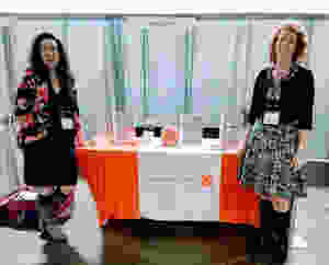 Cassy Beckman, COO and Jolene Thibedeau Boyd, CEO stand next to an orange exhibit table displaying information on Stop Making It Weird at the 2019 Forum on Workplace Inclusion in Minneapolis, MN.