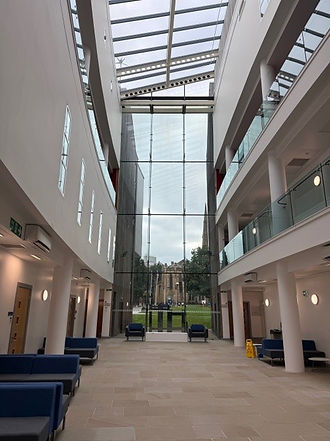 A landscape featuring the interior of the Richard Steinitz Building, the music technology centre at the University of Huddersfield. The scene is of a bright, spacious atrium, with light stone flooring and white walls. Blue and grey cushioned seats are arranged around the room’s perimeter near marked doors. A picturesque floor-to-ceiling glass wall at the far end of the atrium provides a view of a church outside, and a glass roof floods the environment with natural light. Walkways lined with automatic doors on the upper levels are visible from the ground floor, and are enclosed with glass balustrades.