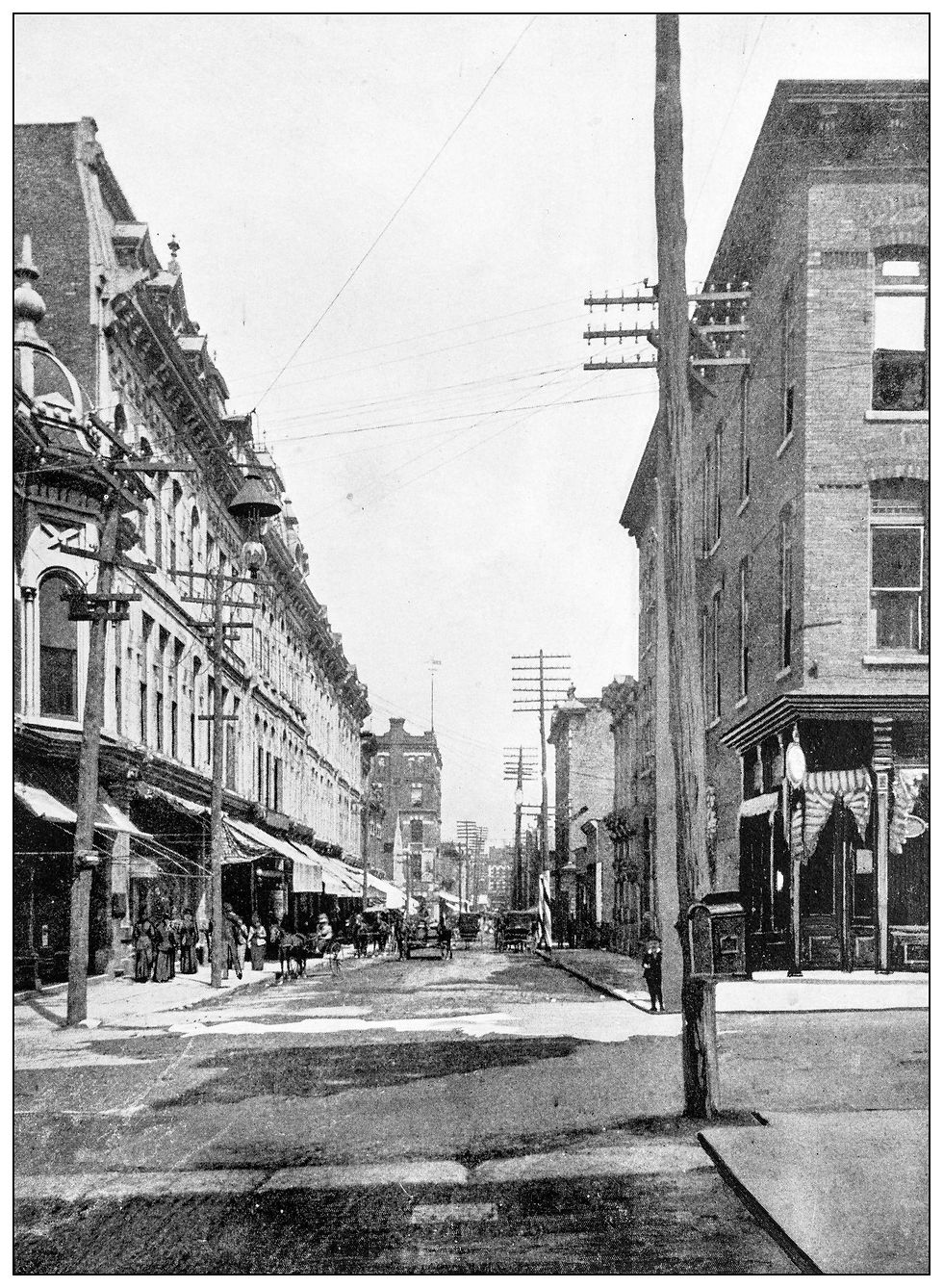 Black-and-white view of a busy old city street with rowhouses, telegraph poles, pedestrians, and a horse-drawn carriage.