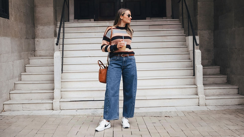 Mujer con suéter de rayas y jeans, sosteniendo bebida. Lleva bolso marrón y gafas de sol. Escalera de fondo. Ambiente casual.