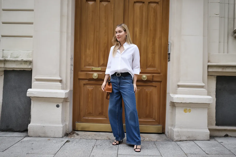 Mujer con camisa blanca y jeans anchos, parada frente a puerta de madera. Lleva bolso marrón y sandalias negras en calle urbana.