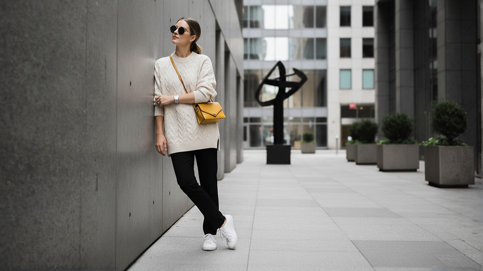 Mujer con gafas de sol apoya en pared, viste suéter beige y bolso amarillo. Fondo urbano de edificios gris oscuro, planta y escultura negra.