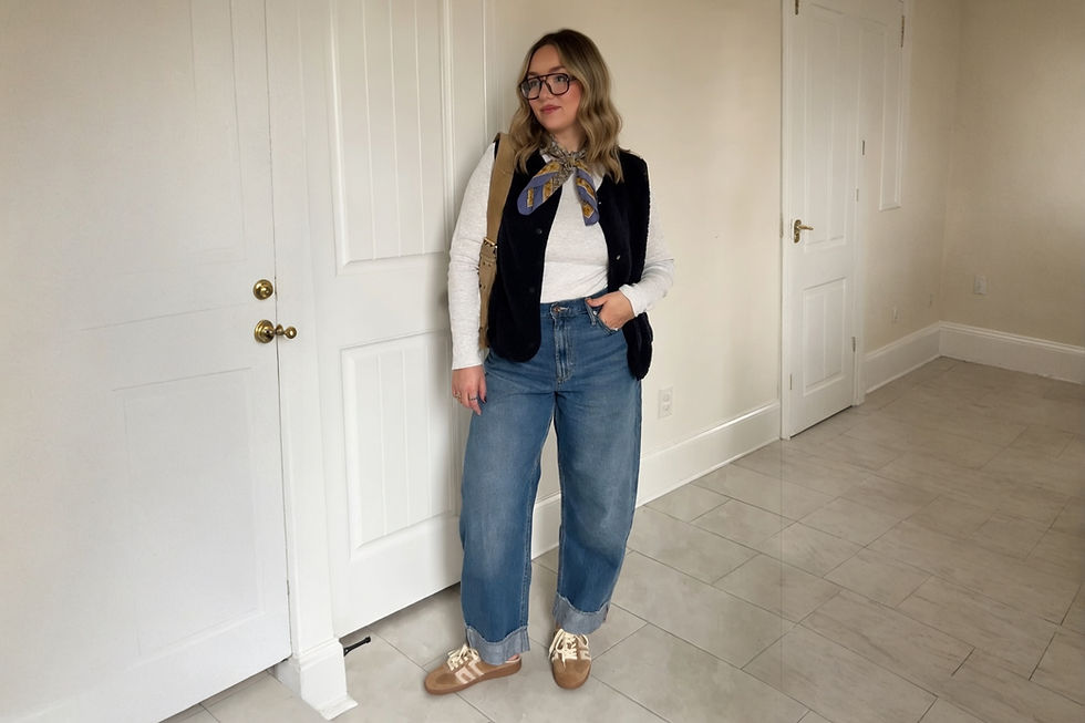 Mujer con gafas, blusa blanca, chaleco negro, jeans y bufanda estampada, posa en habitación de suelo gris y puertas blancas. Relajada.