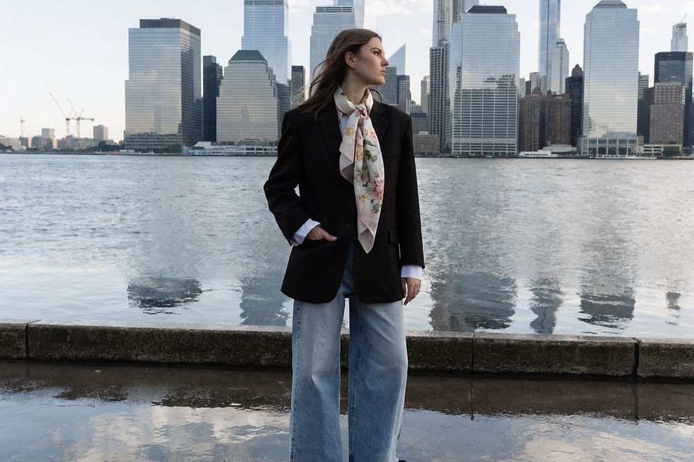 Mujer mira al horizonte junto a un río, viste chaqueta negra y pantalones anchos. Al fondo, rascacielos reflejados en el agua.