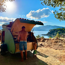 The man stands in an outdoor kitchen, cooking under the open sky. He enjoys the view of the sea while he cooks.