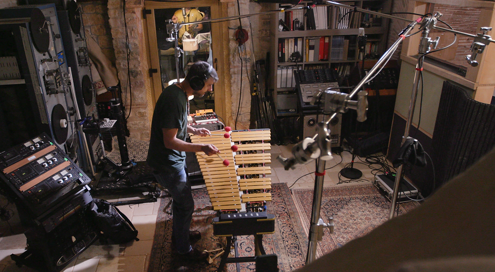 Un'ampia veduta dall'alto all'interno dello studio registrazione Nightingale Studios. Al centro, Alessandro Bianchini è in piedi mentre suona un vibrafono con i battenti a testa rossa, circondato da un'articolata serie di aste microfoniche e giraffe. Sulla sinistra spiccano imponenti registratori a nastro a bobina aperta montati su rack verticali, con strumentazione analogica retrò retroilluminata. Sullo sfondo si notano una porta a vetri che rivela un'altra stanza di ripresa, pareti in pietra, tappeti persiani e una scaffalatura piena di attrezzature audio e nastri.