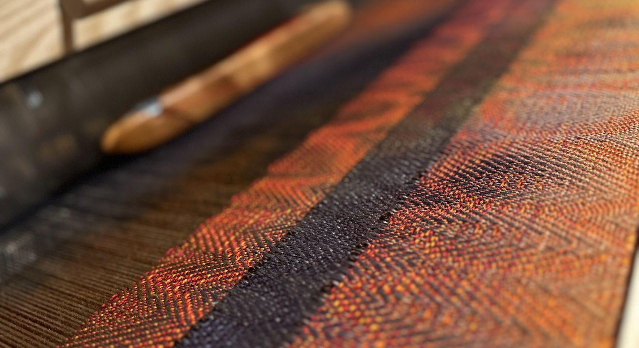 Orange, Yellow, Red and Eggplant colored woven textile on a loom.