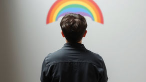Person in black shirt facing a rainbow painted on a plain white wall, conveying a sense of contemplation and hope.
