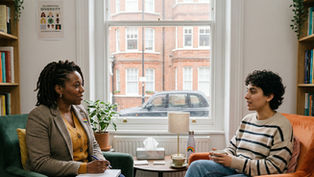 Two people sit in an office, engaged in conversation. Books and plants fill the shelves, with a sign that reads "Celebrating Diversity".