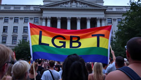 Crowd holding a rainbow LGBT flag in front of a historic building with a dome. The atmosphere is lively and supportive.