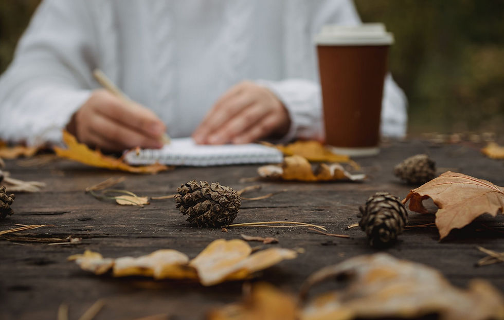 Person writing in a notebook at an outdoor table with fallen leaves and a pinecone. A coffee cup is beside them, creating an autumnal mood.