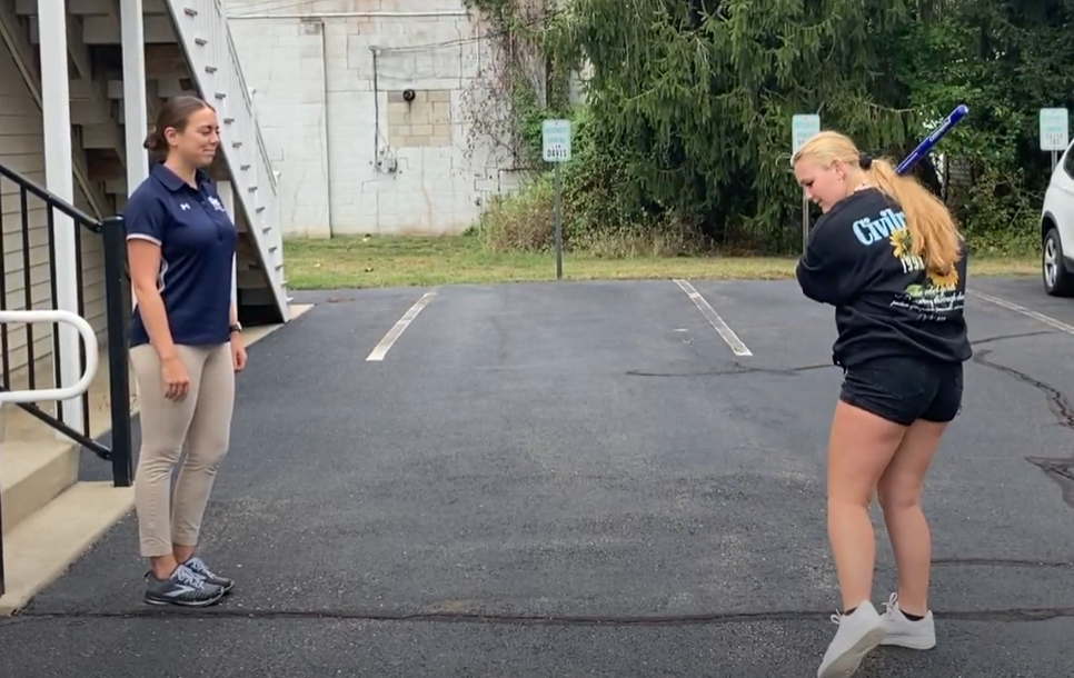 Athlete practicing a softball swing in a parking lot while a coach observes, emphasizing the importance of body balance and symmetry in optimizing swing performance.