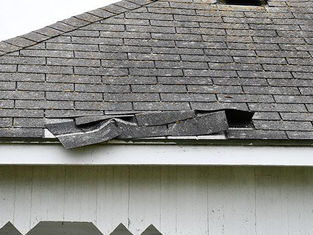 Gray shingled roof with damaged, buckled section. White house exterior. Overcast sky. No text visible. Mood suggests wear or disrepair.