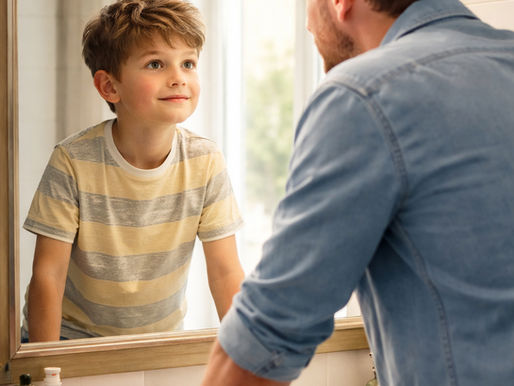 Man in denim shirt leans on a bathroom sink, looking at his son in a striped shirt through the mirror. Bright, warm lighting fills the room.