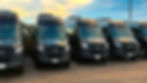 Five black Mercedes-Benz vans parked in a row under a blue sky with clouds. License plates visible. Calm, organized setting.