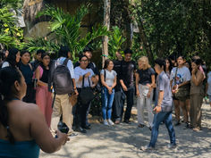 A group of people standing outdoors near a thatched-roof hut, surrounded by lush green foliage, in a sunny setting.