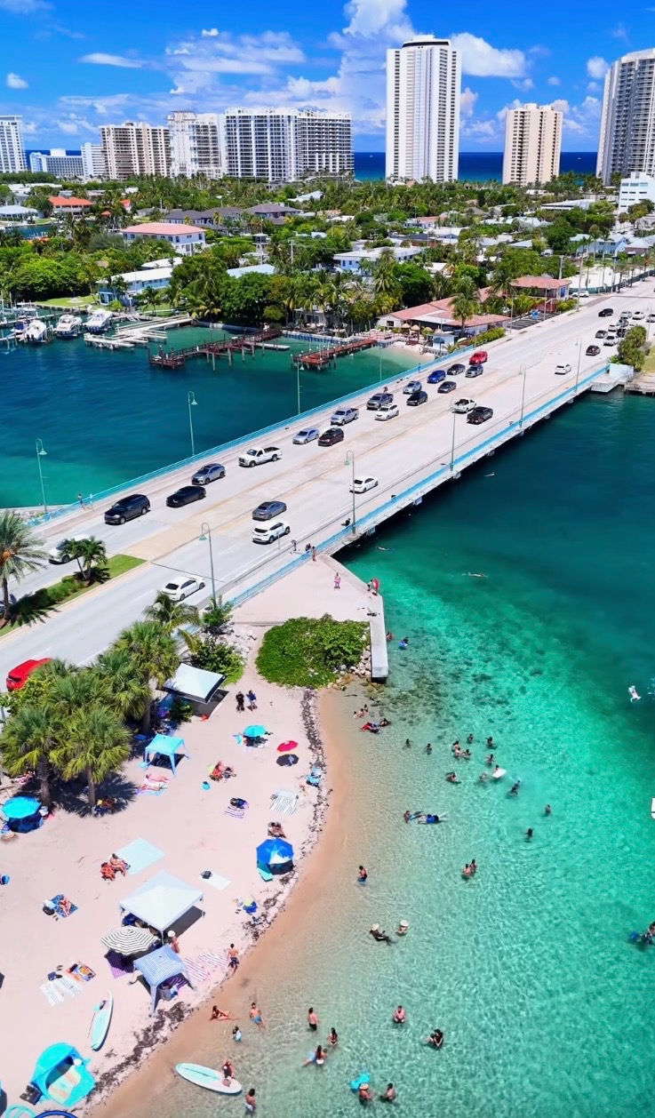Beach scene with people swimming in turquoise water. Nearby, umbrellas and palm trees. Bridge with cars above. Cityscape in the background.