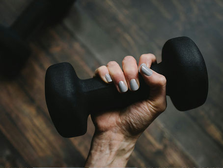 A close-up of a hand with light gray painted nails gripping a black dumbbell over a wooden floor.