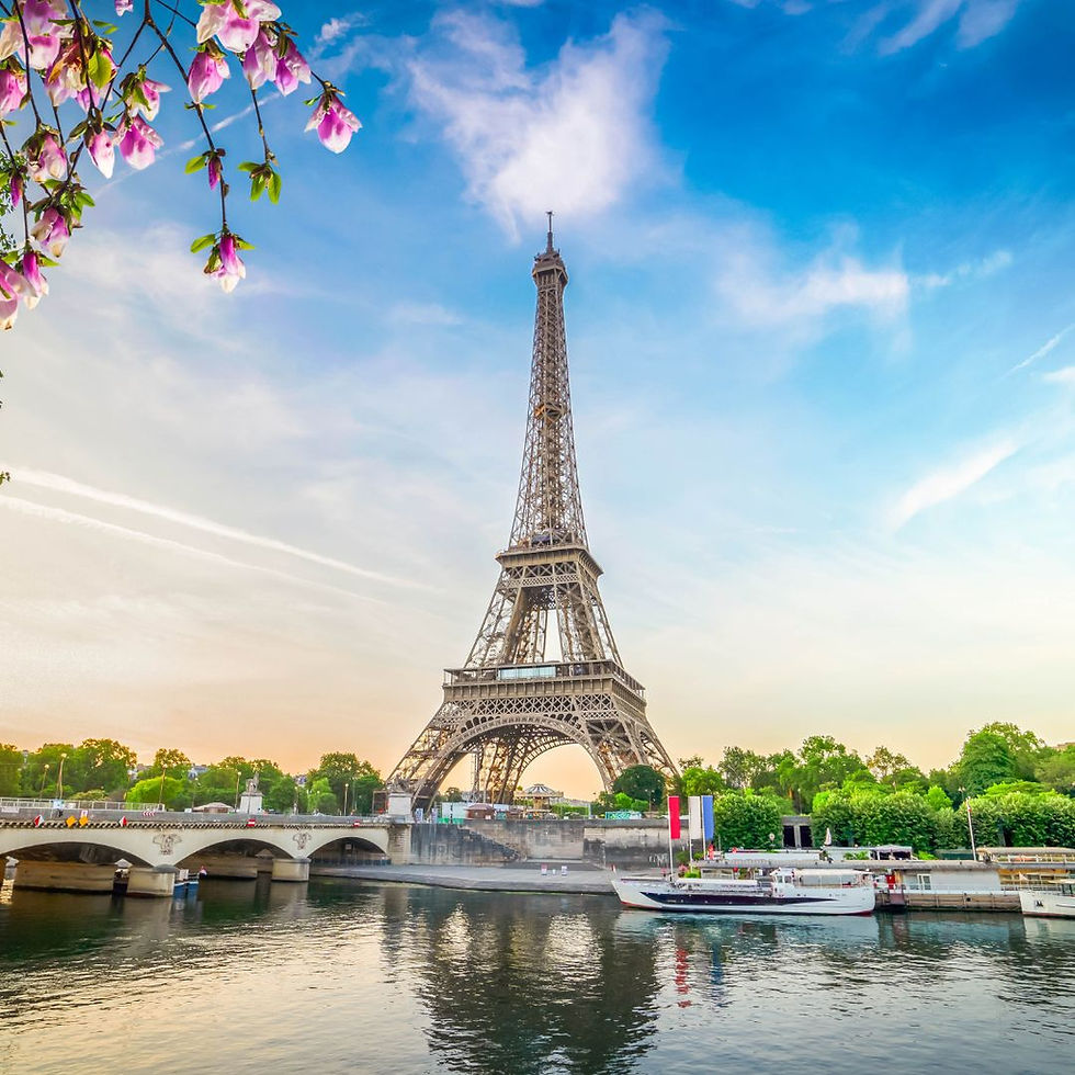 The Eiffel Tower framed by pink spring blossoms in the upper corner, with the Seine River, a stone bridge, and tour boats in the foreground during a soft sunset.