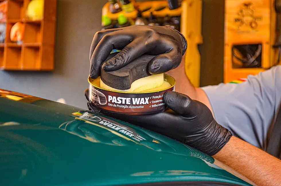 Uma pessoa realizando a aplicação de Cera em uma lataria de carro verde, ele segura a cera com uma mão e a esponja aplicadora em outra