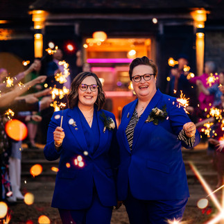 Newly weds in matching blue suits walk through a sparkler exit at their alternative wedding, celebrating a joyful LGBTQ+ ceremony with guests cheering behind them.