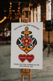 tattoo inspired wedding sign on an easel stood in a large mill venue with lights behind