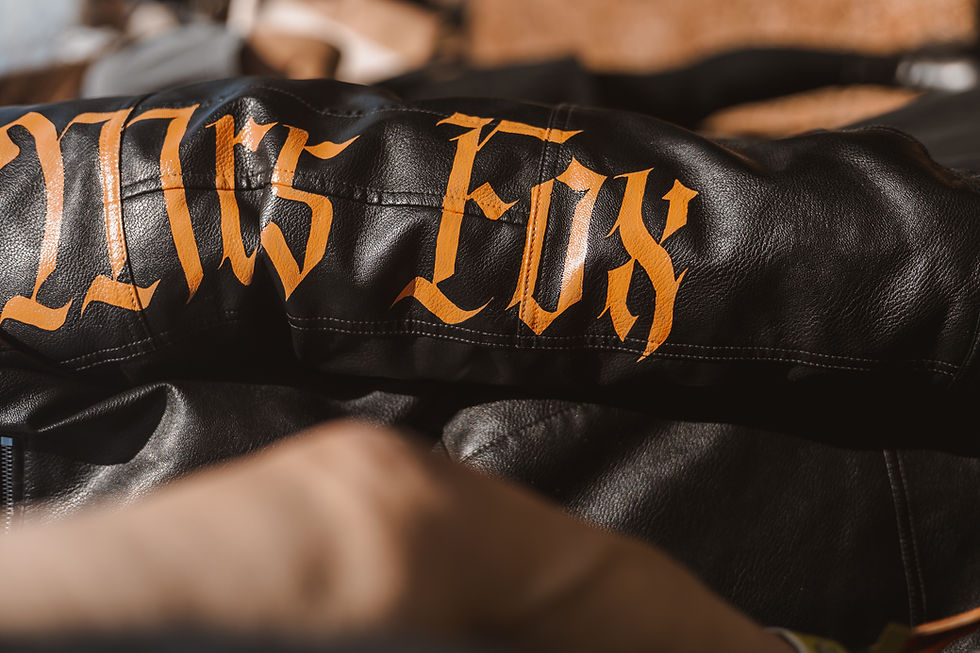 Close-up of a black leather jacket with orange gothic lettering, blurred behind another jacket in warm light.