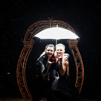 photo in the dark of a couple under a umberella that is lit up, one is sticking their tongue out and they are both doing horns at the camera