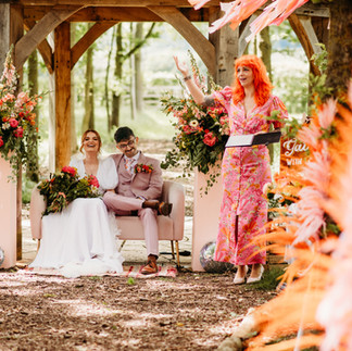  a couple sat under a wooden cover in their wedding outfits, a white dress and a pink suit, with a celebrant in a pink dress with bright orange hair laughing and smiling