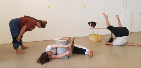 Three performers engaged in floor-based movement, interacting with everyday objects like a chair, plank, and stool .
