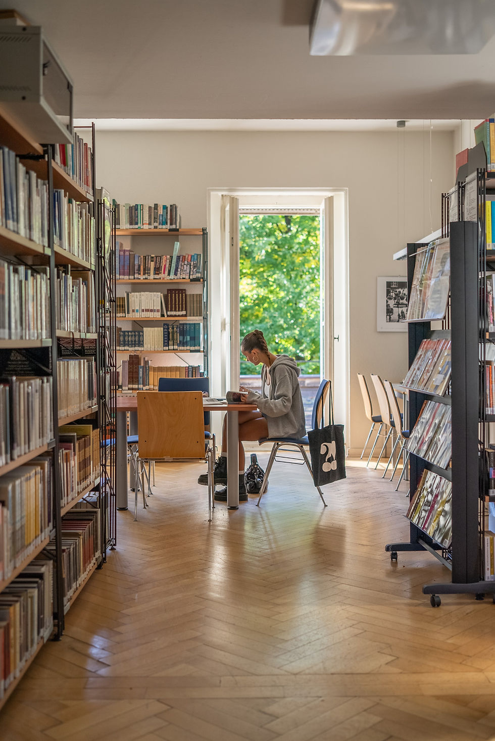 Young woman reads at a library table between bookshelves, with sunlight and green trees visible through an open door.