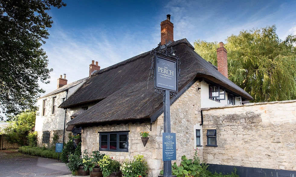 Charming stone pub with thatched roof under blue sky, surrounded by greenery. Sign reads "The Perch, Binsey Village, Oxford."