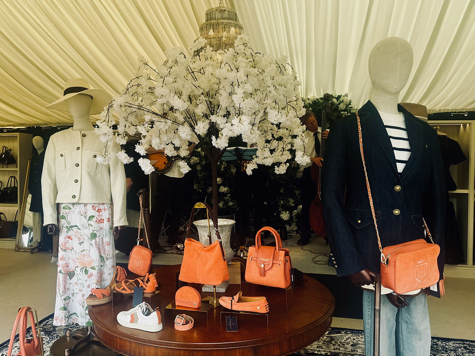 Mannequins in chic outfits under a white tent, displaying orange accessories. Elegant decor includes a white blossom tree and a chandelier.
