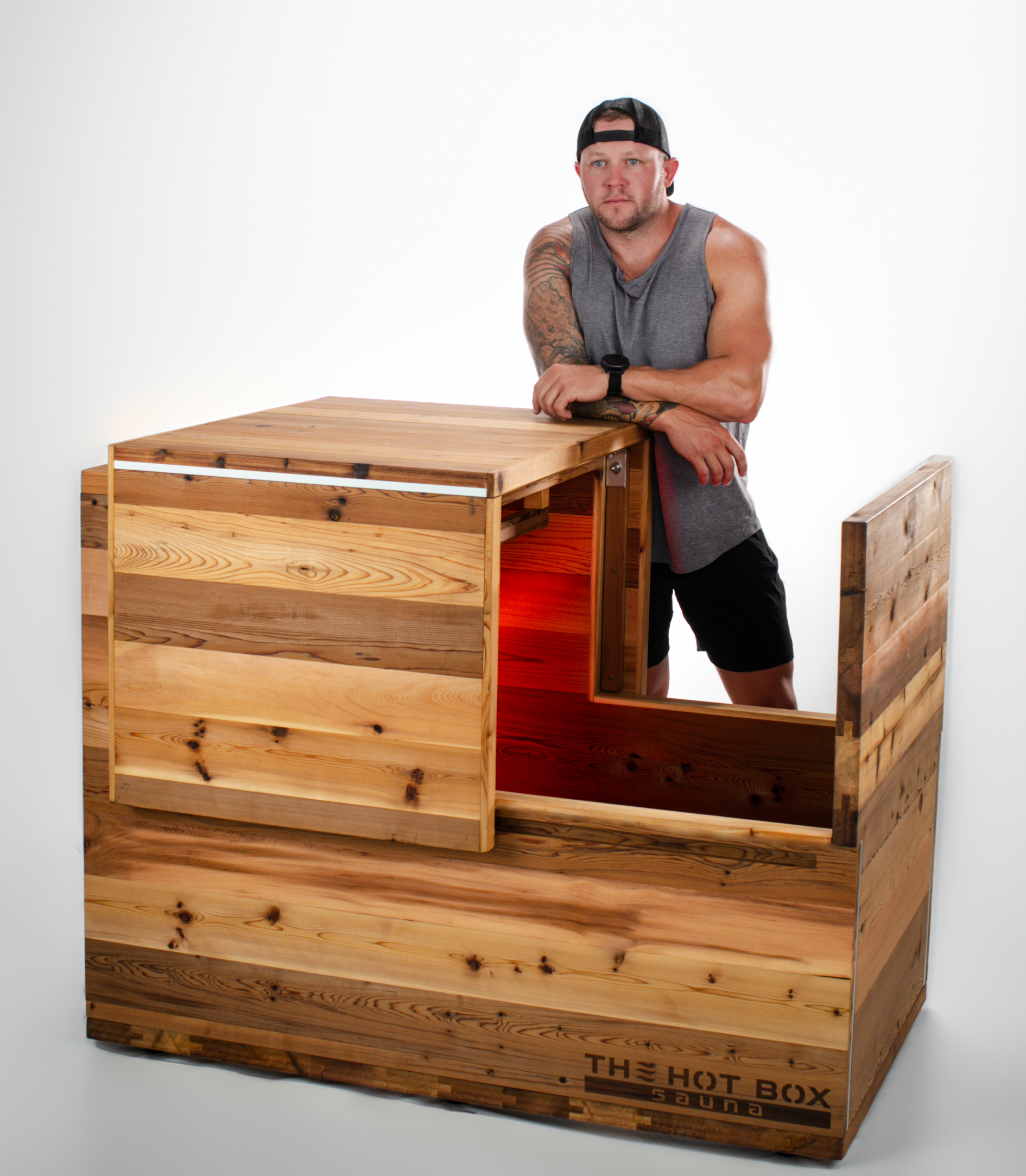 A man leaning against The Hot Box Sauna Standard with the lid raised, showing the bench and interior of the cabin
