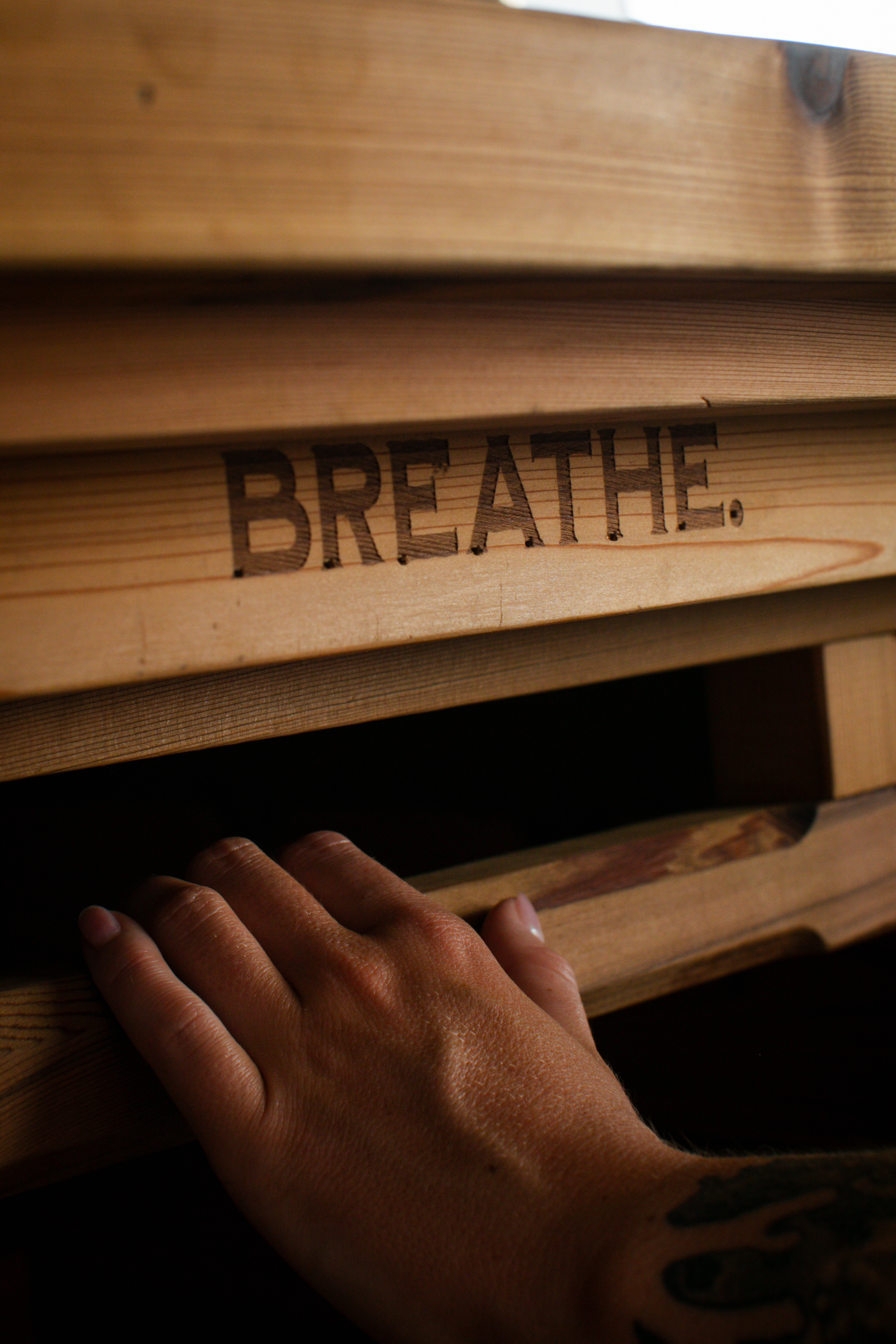 A hand resting beside the word BREATHE engraved into the wood bench of a Hot Box Sauna