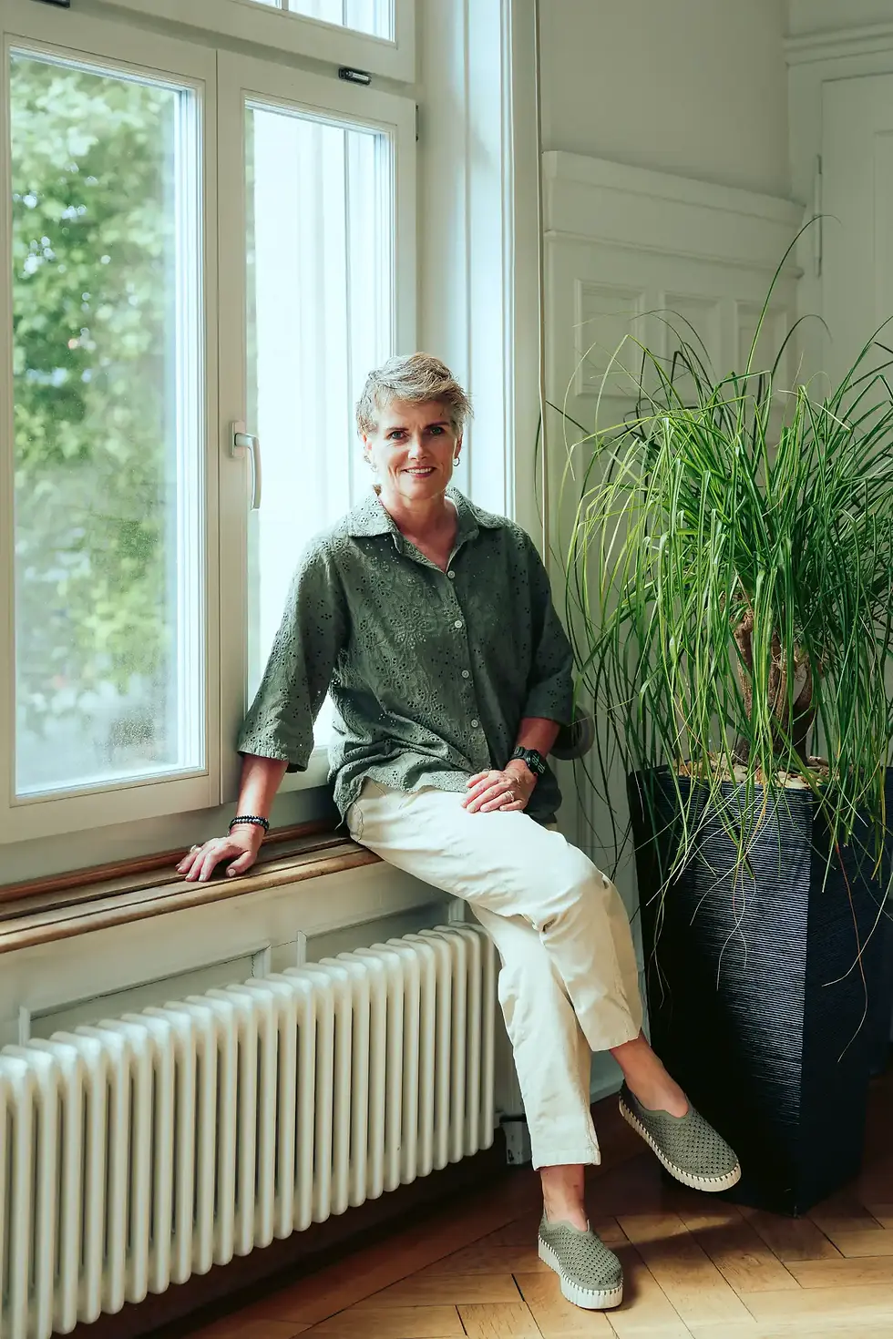 Porträt von Esther Müller, Ergotherapeutin, sitzend am Fenster neben einer großen Zimmerpflanze.
