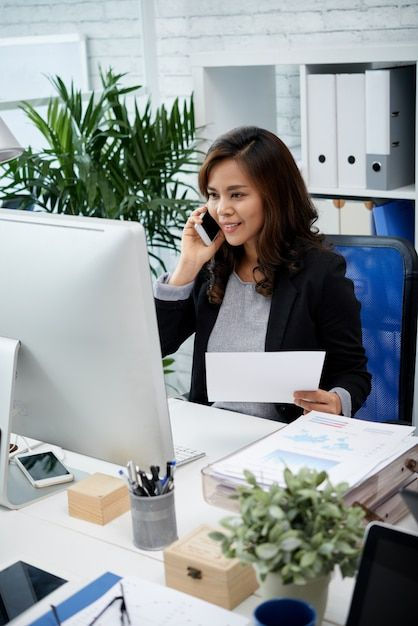 Asian business lady sitting in office, holding document and talking on mobile phone _ Free