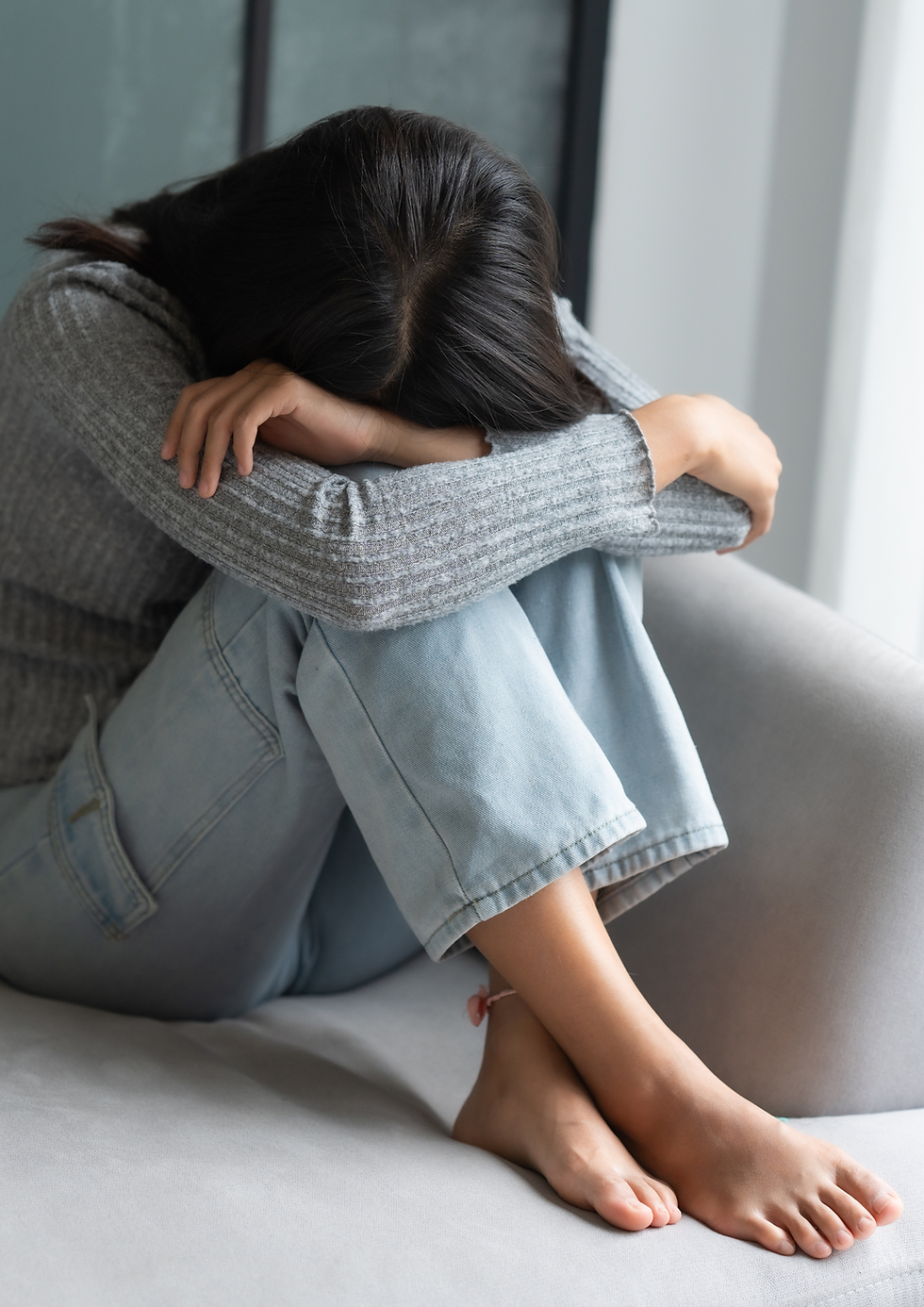 Mulher em jeans e suéter cinza sentada no sofá, cabeça nos braços. Ambiente claro e neutro. Expressa tristeza ou reflexão.