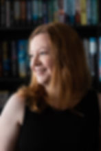 Smiling woman with red hair in a black top, looking to the side against a background of colorful bookshelves. Bright lighting.