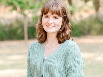 A white, 20-something woman with light brown hair posing for a headshot in front of a background of trees.