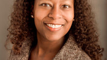 A woman of color with brown hair, skin, and eyes poses for a headshot in front of a brown background.